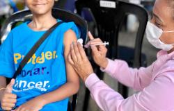 Criança sorridente, de camiseta azul, recebe uma injeção no braço enquanto faz sinal de positivo com a mão. Uma profissional de saúde, de máscara e blusa rosa, aplica a vacina. Cena de vacinação ao ar livre.