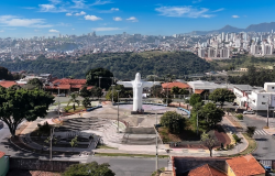 Praça no Barreiro vista de cima. Cristo com os braços abertos no centro da praça, ao seu redor algumas árvores, casas e ruas. No plano de fundo a cidade de Belo Horizonte. 