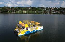 Barco turístico, nas cores branco, azul e amarelo, navegando na Lagoa da Pampulha, mostrando a orla ao fundo