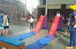 Crianças brincam em dois escorregadores em quadra poliesportiva com trave, acompanhadas por monitor. 
