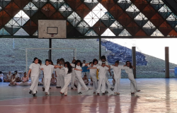 Mais de quinze crianças de branco dançam de maneira coordenada em ginásio. 