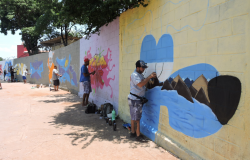 Cinco jovens grafitam diferentes partes de um muro, cada uma delas com uma cor de fundo, durante o dia. 