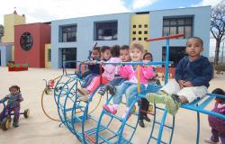 Sete crianças brincam em playground a frente de Unidade de Municipal de Educação Infantil, durante o dia. 