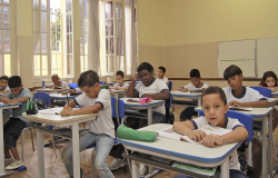 Nove crianças em sala de aula, sentadas em carteiras, durante o dia. 