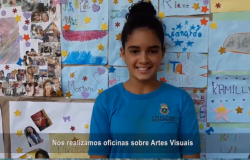 Estudante à frente de uma painel feito por alunos com legenda que diz: "Nós realizamos oficinas sobre Artes Visuais", cena de vídeo documentário Amabilidade da E. M. Santos Dumond. 