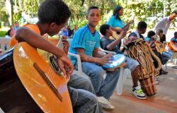 Menino toca violão, acompanhado de outras crianças que tocam pandeiro, tambor e violões, em local aberto, durante o dia. 