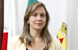 Roberta Rodrigues Martins Vieira Mulher posa em frente à bandeira de Belo Horizonte e do Estado de Minas Gerais, usando casaco amarelo