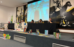 fotografia colorida de uma mesa, composta por cinco professores(as). A mesa é decorada com livros e uma faixa, na qual está escrito: “Somos todos iguais nas diferenças!”