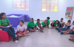 Na sala sensorial, com uma piscina de bolinhas no fundo, à esquerda, e um mural sensorial na parede à direita, a foto mostra uma meia lua formada por estudantes. Enquanto os(as) outros(as) se sentam no chão, Melissa está sentada em um pufe. 