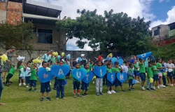 Crianças pequenas uniformizadas estão reunidas em um gramado, durante uma passeata ao ar livre. Na frente do grupo, algumas seguram grandes corações azuis de cartolina com uma letra que, ao se juntarem, formam a palavra "autismo". Atrás delas, outras crianças e pessoas adultas seguram enfeites coloridos, como pompons e flores de papel. Ao fundo, há árvores, um muro de cimento e algumas casas.