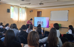 Na imagem apresentada, as alunas estão em um local fechado, assistindo a uma apresentação de slides, projetada em um telão, onde está escrito “Não sou mais uma menina... E agora?” Elas estão em uma sala com paredes pintadas em cor de laranja suave. À esquerda, há uma janela com cortinas brancas e, a seu lado, está um ventilador. No teto também há um ventilador. Ao fundo do palco, está afixado um quadro branco. Em frente ao telão, uma mulher segura um microfone.