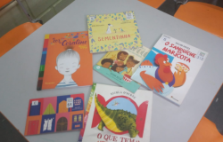 #paratodosverem: em uma mesa cinza cercada por cadeiras laranjas, estão dispostos seis livros infantis coloridos. Entre eles, “A cor de Coraline”, “Sementinha”, “O sanduíche da Maricota”, “O que tem atrás da porta?”, “O que tem atrás do medo?” e “A casa e o seu dono”. As capas trazem ilustrações vibrantes de animais, pessoas e cenários lúdicos.