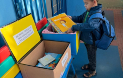 #paratodosverem: Um menino de mochila azul e casaco observa um livro infantil intitulado “Minha Pele” diante de caixas de papelão sobre um banco colorido. As caixas têm placas com os dizeres “PROJETO: TROQUE E DOE UM LIVRO DE LITERATURA INFANTIL” e “TROQUE”. Dentro delas há outros livros disponíveis para troca.