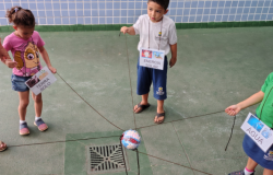 #paratodosverem: Três crianças participam de uma atividade ao ar livre, segurando cordões que se cruzam no centro, onde há uma bola representando o planeta Terra. Cada uma usa um crachá com palavras: “TERRA SOLO”, “ENERGIA SOLAR” e “ÁGUA”, simbolizando os elementos da natureza. O fundo é um muro azul quadriculado.