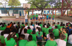 #paratodosverem: em uma quadra escolar coberta, diversas crianças sentadas, vestidas com uniforme verde, assistem a uma apresentação. À frente, uma profissional de jaleco branco está ao lado de uma mesa com materiais e de um grupo de estudantes. 
