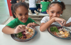 #paratodosverem: a imagem mostra duas meninas sorrindo enquanto fazem uma refeição em um ambiente escolar. Elas estão sentadas à mesa com pratos de comida equilibrada, contendo arroz, feijão, vegetais como couve e beterraba, além de pedaços de frango. A menina à esquerda veste uma camiseta verde, enquanto a menina à direita usa uma blusa branca e tem um laço azul no cabelo. Ao fundo, outras crianças também estão se alimentando.