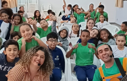 #paratodosverem: Em um auditório com cadeiras brancas, um grande grupo de crianças e adultos está posando para a foto. A maioria sorri, faz "joinha" ou levanta a mão, demonstrando entusiasmo. Em primeiro plano, uma mulher de cabelo cacheado e um homem tocando violão olham para a câmera. A cena é alegre e passa a sensação de um evento ou apresentação de sucesso.