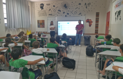 #paratodosverem: a imagem mostra uma sala de aula, com decoração temática da cultura pop. Os(as) alunos(as) estão uniformizados(as) de verde e branco e sentados em carteiras escolares organizadas em fileiras. As crianças estão atentas à apresentação feita por profissionais à frente da sala. Uma mulher, vestindo blusa listrada e calça preta, está com os braços cruzados ao lado de um homem de camiseta vermelha e jeans, que parece estar falando. Atrás de ambos, projeta-se uma apresentação.