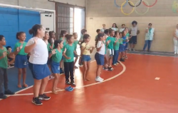 #paratodosverem: em uma quadra, duas fileiras de estudantes se apresentam para o restante da escola. Atrás delas, há uma parede pintada de azul, em que outras crianças estão encostadas, assistindo, sentadas, à apresentação dos(as) colegas. À direita, em frente a uma parede com desenhos coloridos, incluindo os anéis olímpicos, pessoas adultas observam a atividade. 