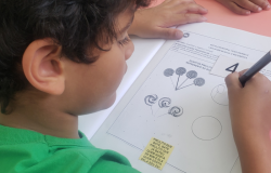 #paratodosverem: na foto, uma criança com camiseta verde realiza uma atividade escolar, que consiste em contar pirulitos desenhados e escrever o número correspondente. A criança segura um lápis e está prestes a escrever. No canto inferior da folha, há a letra da cantiga "Pirulito que bate, bate / Pirulito que já bateu / Quem gosta dela sou eu!"