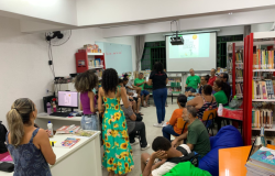 #paratodosverem: na imagem, pessoas adultas participam de uma roda de conversa em uma biblioteca escolar. A maioria está sentada em cadeiras e pufes coloridos. Ao fundo, uma mulher de camiseta preta conduz a atividade, com um slide projetado no quadro em que está escrito “O que você faz com uma ideia?” Estantes com livros, mesas e um computador compõem o ambiente. Algumas pessoas de pé acompanham a conversa.