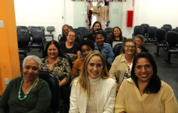 #paratodosverem: na imagem, doze mulheres estão reunidas em um auditório, sentadas em cadeiras de auditório e sorrindo. O auditório tem paredes brancas, cadeiras escuras e uma porta de vidro, ao fundo, decorada com bandeirinhas coloridas. 