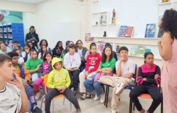 #paratodosverem: a imagem mostra uma roda de conversa em uma biblioteca. Um grupo de crianças e adolescentes está sentado, prestando atenção em uma mulher que fala de pé, à frente do grupo. Ao fundo, há estantes com livros, cartazes e decorações. 