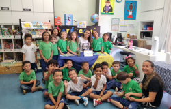 #paratodosverem: foto de uma turma de crianças pequenas uniformizadas em uma biblioteca. Elas estão reunidas em volta de uma mesa decorada com tecido azul e amarelo, acompanhadas por duas educadoras. No fundo, há prateleiras com livros e desenhos coloridos nas paredes.