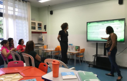 #paratodosverem: na foto, há quatro mulheres, à esquerda, sentadas em cadeiras plásticas brancas, enquanto duas estão de pé, à frente da sala. Uma destas aponta para uma tela interativa onde está sendo exibido um slide intitulado "Impactos na vida social". A sala tem paredes e cortinas brancas, armários com portas de vidro, além de mesas e cadeiras nas cores laranja, azul e vermelha.