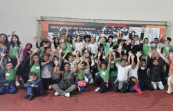 #paratodosverem: a foto mostra um grupo grande de crianças e pessoas adultas reunidas em um ambiente interno. A maioria está sorrindo e com os braços erguidos. As crianças usam uniformes escolares verdes ou brancos e portam crachás de identificação. Ao fundo, há um painel decorativo com o texto "Sertão mineiro. Ser tão mineiro."