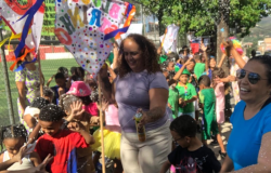 #paratodosverem: fotografia colorida de pessoas adultas e de crianças fantasiadas, celebrando o Carnaval em um espaço aberto, com uma árvore ao fundo, à direita, e grades separando-as de uma quadra aberta, à esquerda.