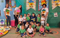 #paratodosverem: A foto mostra um grupo de crianças pequenas sentadas e em pé no pátio escolar, acompanhadas por quatro mulheres. O fundo é decorado com bandeiras coloridas ilustradas com personagens sorridentes e os nomes das turmas, como “Turma do Martelo” e “Turma do Trenzinho”. Há também trabalhos manuais, desenhos e almofadas estampadas com as mesmas figuras. Em meio as crianças, agachada com uma blusa azul, está a artista Raquel Bolinho.