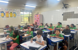 A imagem mostra uma sala de aula com várias crianças uniformizadas sentadas em suas carteiras. Elas estão fazendo uma atividade com papel e lápis. Há um armário rosa ao fundo, decorado com borboletas coloridas, e cartazes educativos na parede ao lado de uma janela gradeada. A iluminação é feita por lâmpadas fluorescentes, e há um ventilador de parede ao fundo, próximo ao teto.