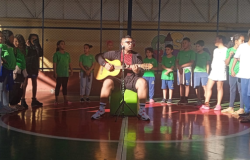 #paratodosverem: em uma quadra, há 15 crianças de pé, dispostas em semicírculo, para apresentação musical. No centro da quadra, um adulto está sentado em um pufe verde, tocando violão com um microfone à sua frente. Os(As) estudantes usam uniformes verdes ou brancos com calças azuis; já o adulto veste uma blusa vermelha, jaqueta e bermuda pretas. 