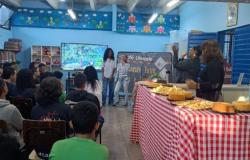 #paratodosverem: a foto mostra o interior de uma biblioteca decorada para o “Café Literário”. Vários(as) estudantes estão sentados(as) de frente para um telão e duas alunas estão, à frente do grupo, apresentando. À direita da imagem, há uma mesa, coberta com toalha xadrez vermelha e branca, repleta de alimentos como bolos, pães de queijo e sucos. Atrás da mesa, duas mulheres assistem às estudantes.
