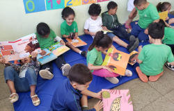 #paratodosverem: Um grupo de crianças, a maioria de uniforme verde, está confortavelmente sentado ou deitado em um tapete azul no chão da sala de aula. Cada criança segura um livro, absorvidas pela leitura, algumas sozinhas e outras próximas.