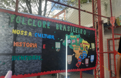 #paratodosverem Painel com o título “Folclore Brasileiro: nossa cultura, história e identidade”, decorado com letras coloridas. No centro, um mapa do Brasil com figuras de personagens folclóricos distribuídos pelas regiões. O cartaz está preso em uma grade de ferro, com bandeirinhas de festa junina ao redor.