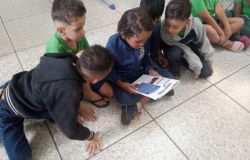 #paratodosverem: Quatro meninos estão sentados no chão de um ambiente escolar. Eles se reúnem em volta de um pequeno jornal ou revista, lendo atentamente. A cena, que mostra o grupo inclinado e focado na leitura, transmite a ideia de um momento de aprendizado e curiosidade compartilhado entre os amigos.