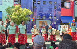 #paratodosverem: Em um palco ao ar livre, um grupo se apresenta. As mulheres vestem blusas verde-água e saias longas vermelhas, enquanto outras tocam instrumentos como sanfona e tambor. Ao fundo, um prédio azul vibrante e um telão exibindo o retrato de uma idosa. A cena é colorida e alegre, com o público assistindo em primeiro plano.