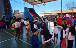 #paratodosverem: A imagem mostra uma quadra esportiva ao ar livre com diversas pessoas, principalmente crianças e adolescentes, participando de atividades. Há brinquedos infláveis coloridos ao fundo, incluindo um escorregador e uma parede de escalada. Algumas pessoas jogam bola e outras observam. Ao fundo, há árvores grandes e construções em tons de vermelho, azul e amarelo.