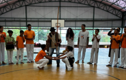#paratodosverem: Um grupo de jovens, vestindo uniformes de cores laranja e branco, está disposto em um semicírculo em um ginásio coberto. No centro, dois participantes estão agachados, tocando as mãos, em um ato de capoeira. Ao fundo, um homem toca um berimbau. A cena ocorre sob um telhado arqueado de metal, com uma tabela de basquete visível.