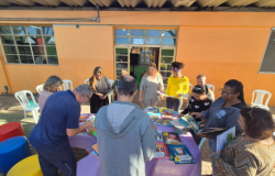 #paratodosverem: Um grupo de adultos está ao redor de uma mesa coberta com toalha roxa, onde há vários livros coloridos expostos. Algumas pessoas folheiam os livros, enquanto outras observam. Ao fundo, vê-se uma parede laranja com janelas de vidro e cadeiras plásticas brancas encostadas.