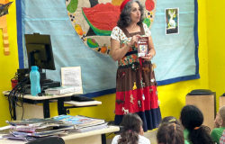 #paratodosverem: a foto mostra uma mulher de vestido colorido e estampado exibindo um livro para um grupo de crianças uniformizadas sentadas no chão de uma sala de aula. Ela está posicionada em frente a um painel azul com um desenho circular ao centro, contendo diversas frutas. Ao lado, há uma mesa com pilhas de livros e outra com um computador. 