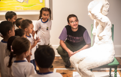 #paratodosverem: grupo de crianças pequenas, uniformizadas, observa atentamente uma escultura branca de figura humana sentada. Ao lado, um educador agachado sorri para os alunos. A cena ocorre dentro de um museu, com obras de arte nas paredes e iluminação suave, criando um ambiente de aprendizado e curiosidade.