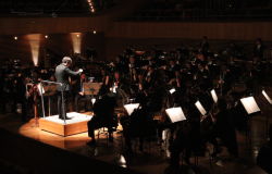 #paratodosverem: a imagem mostra a Orquestra durante a apresentação na Sala Minas Gerais. O maestro está em pé, ao centro, em uma pequena estrutura elevada, regendo, com movimentos das mãos, músicos e musicistas, que estão organizados(as) no palco, conforme os instrumentos: cordas, à frente; sopros, ao centro, e percussão, ao fundo. Luzes incidem sobre o maestro e sobre as partituras. 