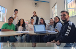 #paratodosverem: Grupo de jovens e um adulto estão sentados e em pé ao redor de uma mesa em sala iluminada, com janelas laterais e um ventilador. Sobre a mesa há um notebook aberto e cadernos. Todos sorriem para a foto, alguns usando moletom, outros camiseta, e uma jovem de touca marrom está em pé ao fundo.