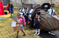 #paratodosverem: Em um jardim, crianças brincam com um avião de papelão feito por elas mesmas. Duas meninas e um menino estão fantasiados de pilotos e tripulação, sorrindo e se divertindo. Uma das garotas, com uma mochila rosa nas costas e um papel nas mãos, olha diretamente para a câmera. A cena transmite a alegria de uma brincadeira cheia de imaginação.