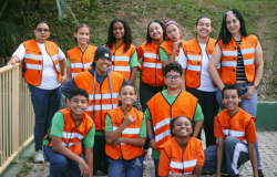 #paratodosverem: a foto mostra um grupo de estudantes com duas educadoras. Todos(as) vestem colete laranja com faixas refletivas brancas e detalhes pretos sobre a blusa. Eles(as) estão ao ar livre e, ao fundo, há um gramado. 