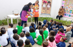 #paratodosverem: Em uma sala de aula, um grupo de adultos está em pé, formando um círculo. Eles seguram fios de barbante, criando uma teia entre si, como parte de uma atividade coletiva. Ao fundo, há cadeiras laranjas encostadas na parede, armários coloridos e materiais pedagógicos. A sala é iluminada por janelas e há um ventilador na parede.
