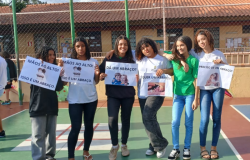 #paratodosverem: a foto mostra um grupo de seis alunas de pé, em uma quadra escolar aberta, sorrindo e segurando cartazes com as mensagens: “Mãos ao alto! Isso é um abraço.”; “Dá um abraço?”; “Quer um abraço?” e “Preciso de um abraço!” Nos cartazes, também há foto de pessoas se abraçando. Ao fundo, aparece a tela que cerca a quadra e uma edificação pintada em amarelo, com portas e janelas marrons.